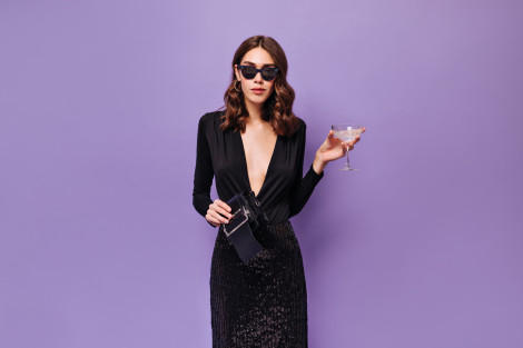 Elegant woman in sunglasses and black dress holds martini glass. Charming lady in festive outfit poses with champagne on purple background.