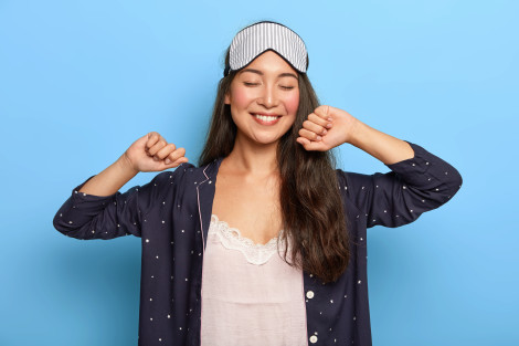 Relaxed joyful Asian woman stretches hands after awakening, rejoices good day, enjoys pleasant sleep, smiles sincerely, wears sleep mask and pyjamas, has happy expression, poses over blue wall