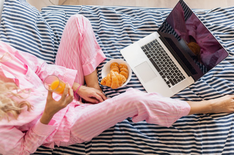 young pretty blond woman in pink pajamas sitting on bed with laptop having breakfast, freelancer at home